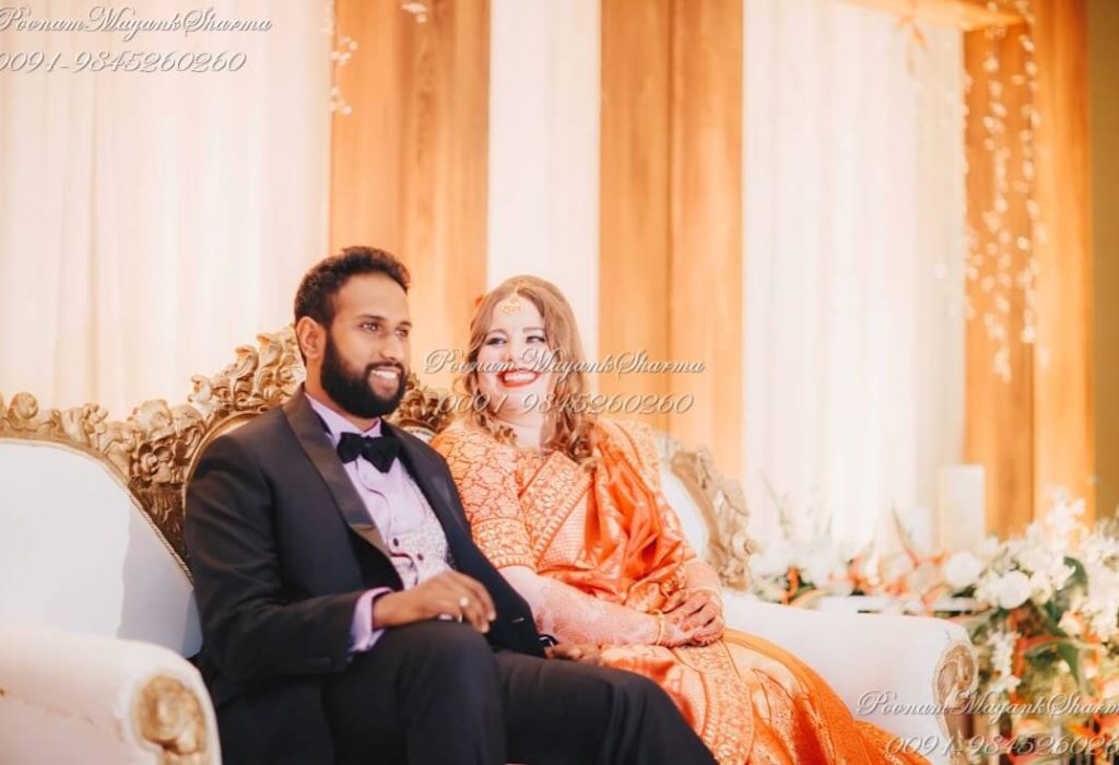 A festive Indo-western wedding organized by Poonam Mayank Sharma. The bride is shining in an orange saree and the groom in a classic tuxedo sits on an elegant royal sofa amidst beautifully decorated floral arrangements.