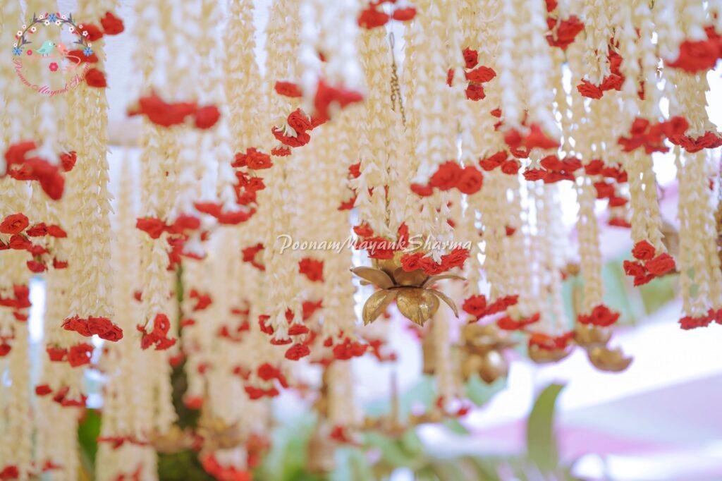 Elegant floral ceiling décor featuring delicate white jasmine and vibrant red roses, adorned with golden lotus-shaped hanging ornaments, designed by Poonam Mayank Sharma, a luxury wedding and event planner in Bangalore, India.